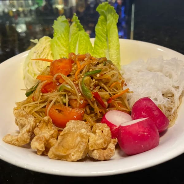 Som Tum (papaya salad) with crispy pork rinds, radishes, lettuce, and vermicelli noodles on a white plate.