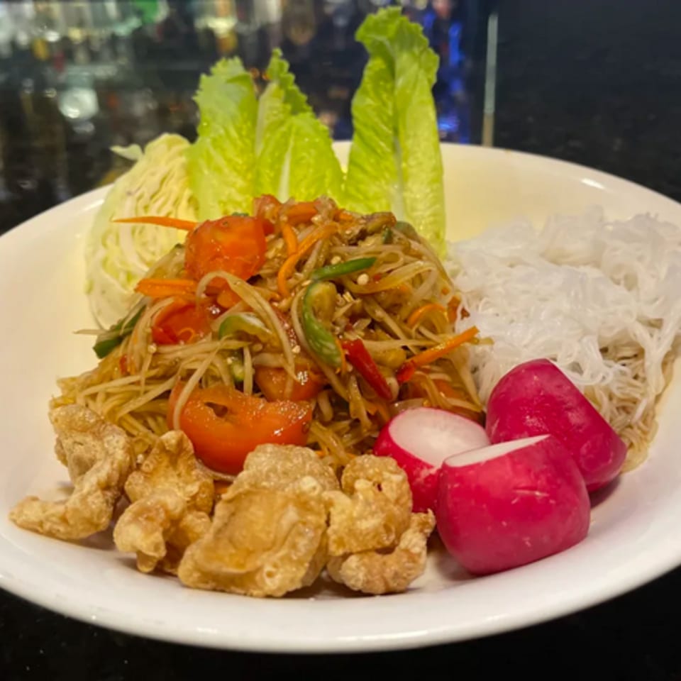 Som Tum (papaya salad) with crispy pork rinds, radishes, lettuce, and vermicelli noodles on a white plate.