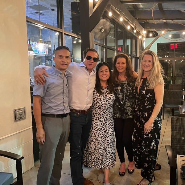 Five smiling people, three women and two men, posing together on a restaurant patio at night.