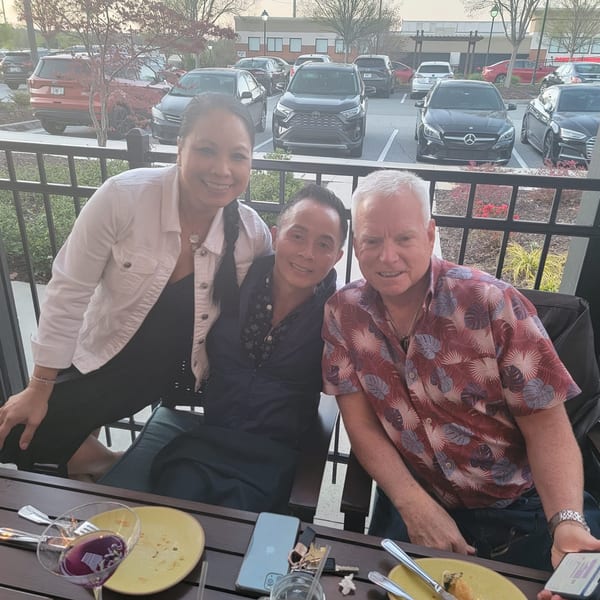 Three smiling people pose for a photo on a restaurant patio with a parking lot in the background.