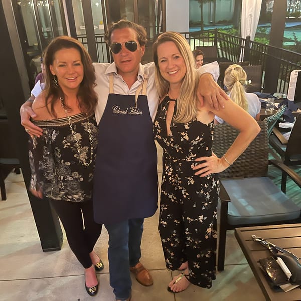 Three smiling people, one wearing a "Colonial Kitchen" apron, posing on a restaurant patio.