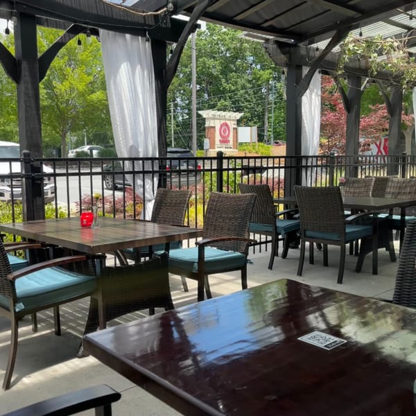 Outdoor patio seating at Colonial Kitchen and Bar, with dark tables, wicker chairs, and white curtains.