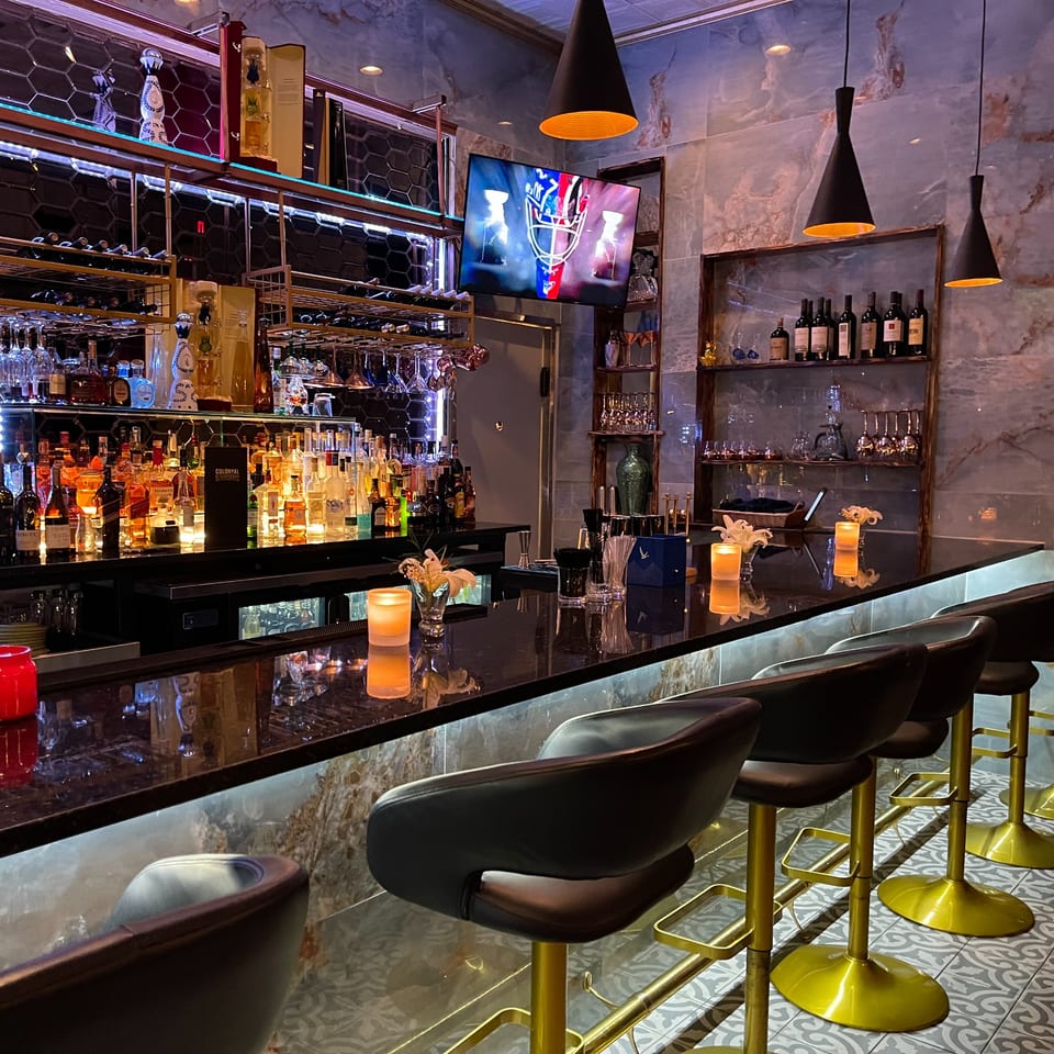 The Colonial Kitchen and Bar features a well-stocked bar with bottles, glasses, and modern lighting.