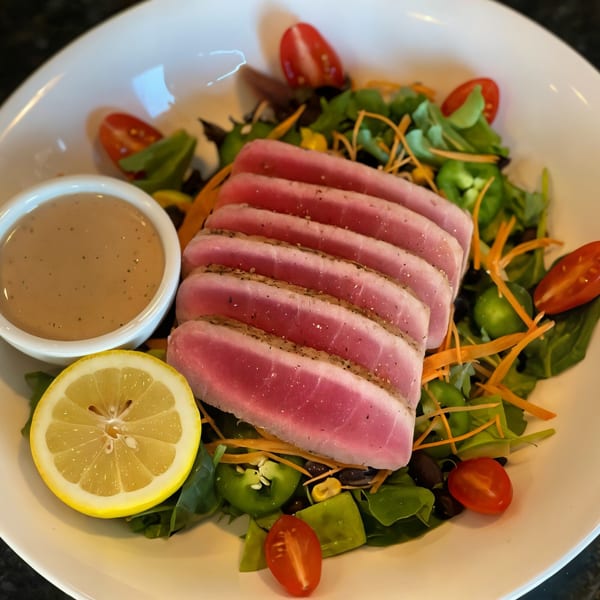 Seared tuna slices on a bed of mixed greens with cherry tomatoes, a lemon wedge, and dressing.