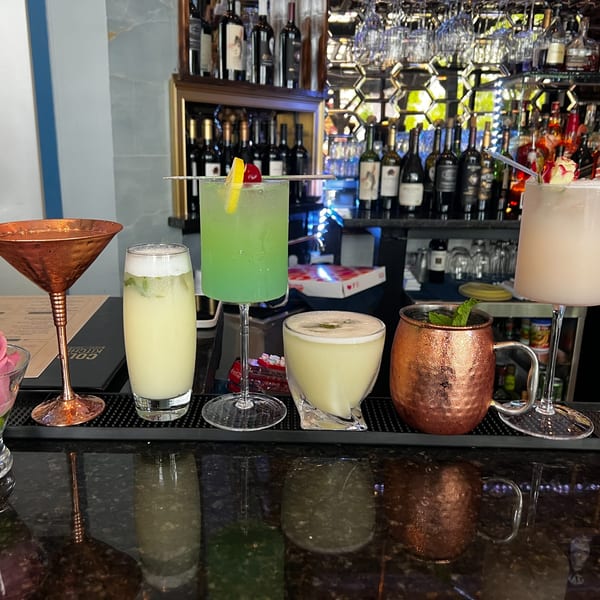 Five colorful cocktails, including green and pale drinks, on a bar counter with bottles in the background.