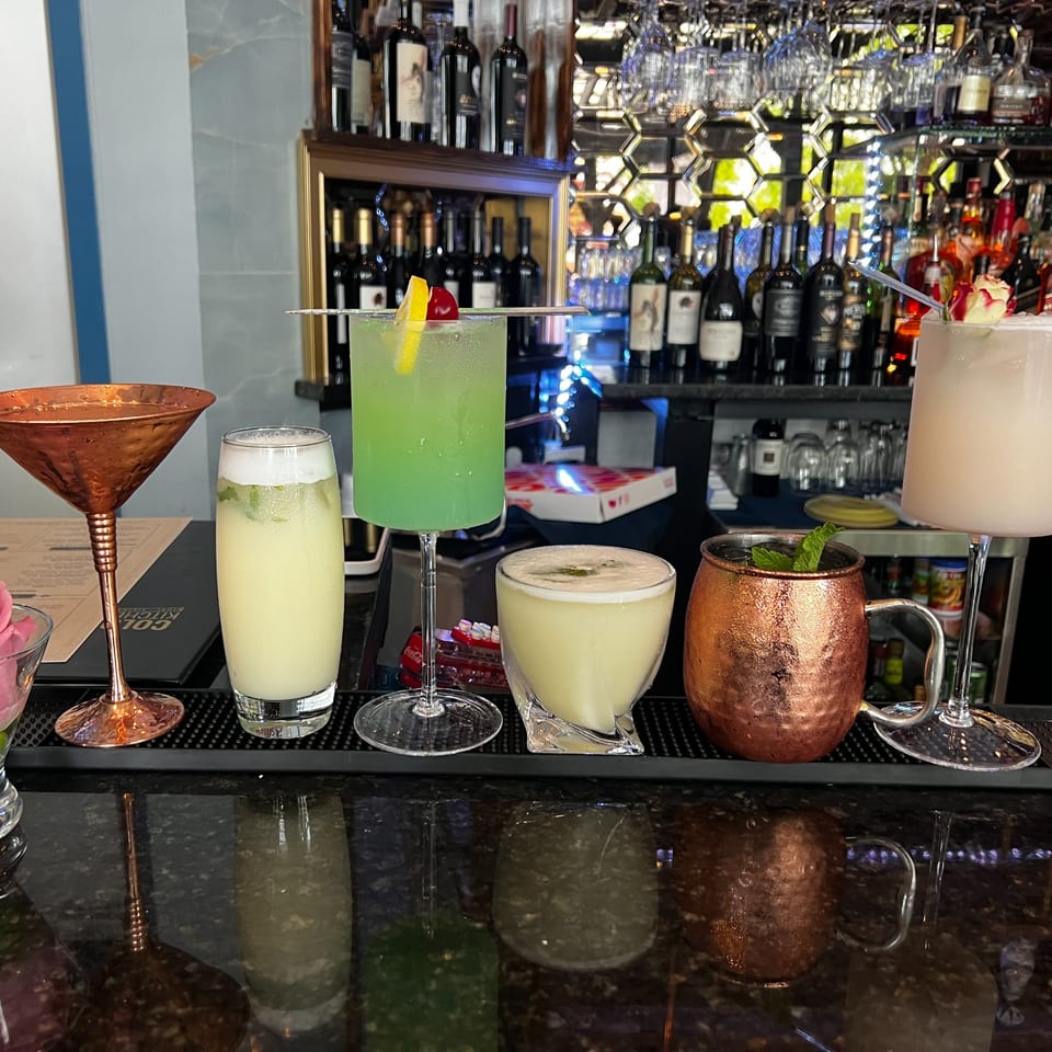 Five colorful cocktails, including green and pale drinks, on a bar counter with bottles in the background.